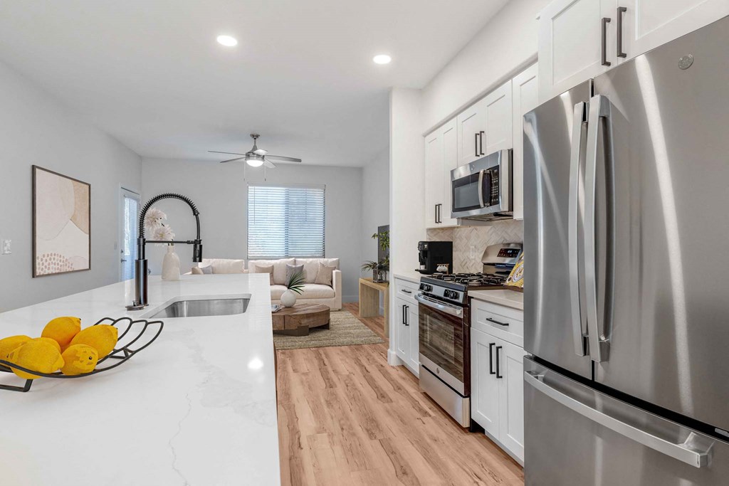 Modern kitchen here at Desert Club Apartments with stainless steel appliances and white cabinets, leading to a cozy living room. Bright space with wood floors and a bowl of lemons on the counter.
