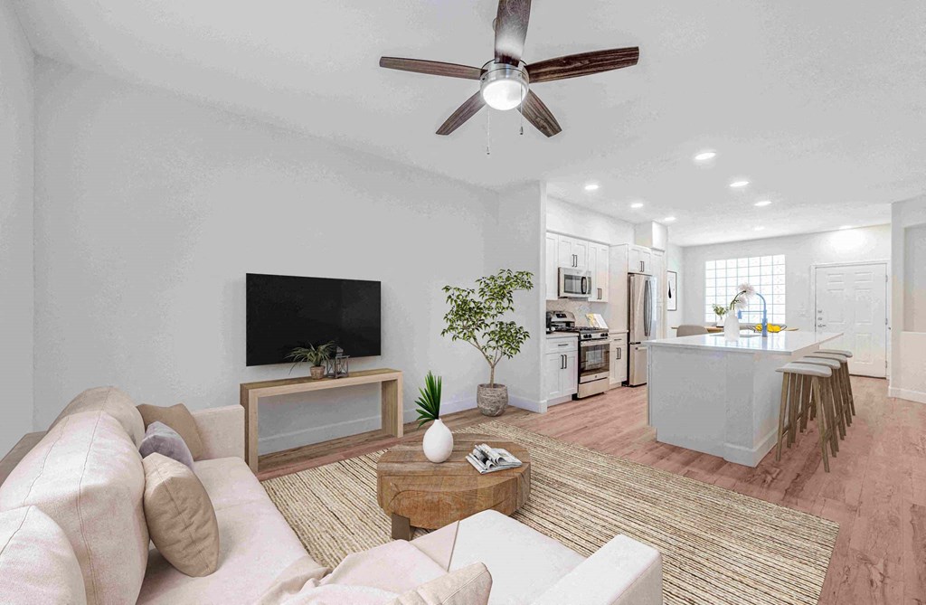 Modern open-plan living room  and kitchen here at Desert Club Apartments with beige sofa, round wooden coffee table, and a TV on a console. Kitchen features white cabinets, stainless steel appliances, island with barstools, and potted plants, creating a cozy, inviting atmosphere.