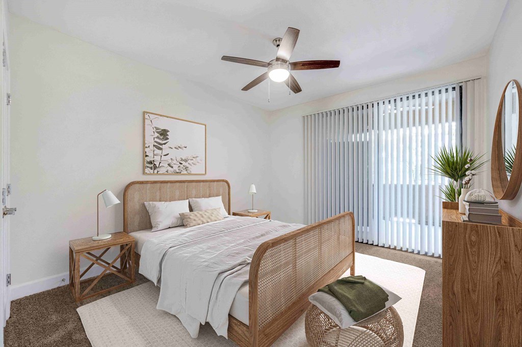 A serene bedroom here at Desert Club Apartments with a rattan bed frame, soft white bedding, and two bedside lamps on wooden tables. A ceiling fan, plant, and blinds enhance the calm vibe.