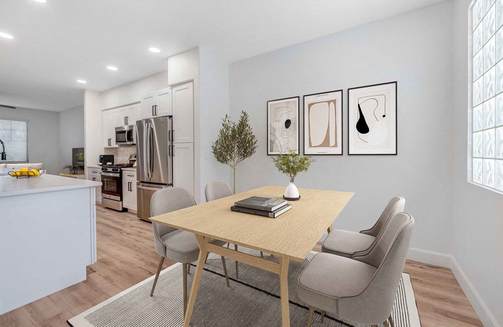 A modern kitchen here at Desert Club Apartments and dining area with wooden flooring. A light wood dining table with four gray chairs sits on a rug. Three abstract artworks adorn the wall, and a plant adds greenery. In the background, stainless steel appliances and white cabinets are visible. Bright and welcoming atmosphere.