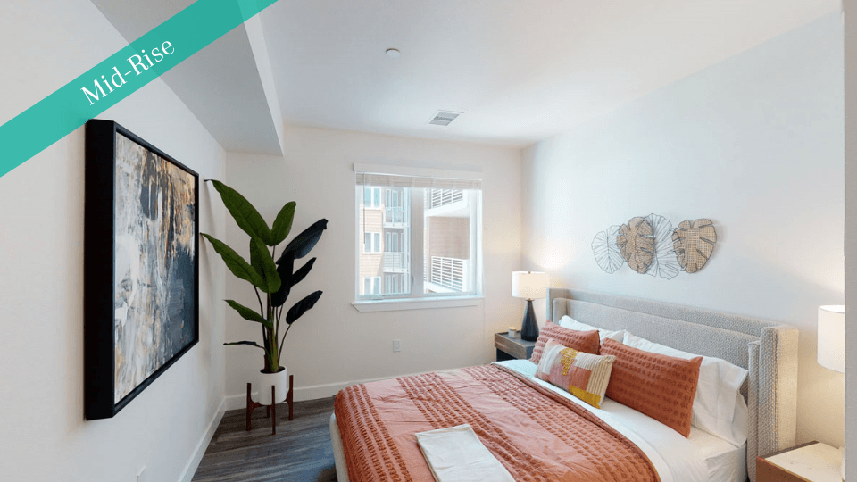 Modern bedroom here at Capitol Towers Luxury Midrise Apartments with a cozy, neatly made bed in warm orange tones. Features a potted plant, abstract wall art, and a calm, inviting atmosphere.