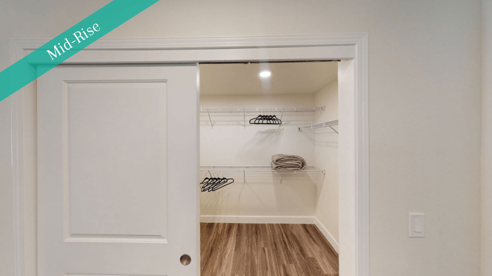 Walk-in closet here at Capitol Towers Luxury Midrise Apartments with wooden flooring, metal shelves, black hangers, and folded towels on the right. It feels modern and organized.