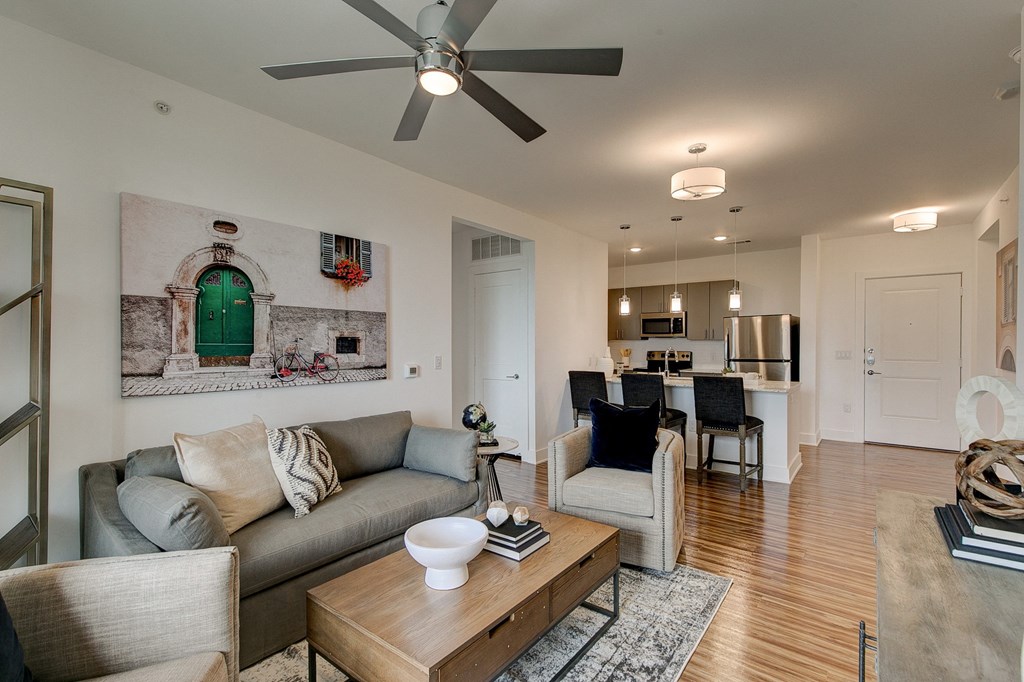 Modern living room here at Florence at the Harbor Apartment Homes with a ceiling fan, gray sofa, and armchair. A large wall art of a green door adds color. Adjacent is a kitchen with pendant lights and a sleek, bright ambiance.