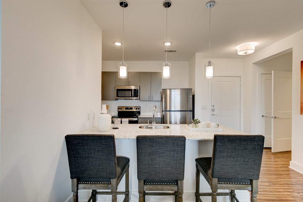 Sleek kitchen here at Florence at the Harbor Apartment Homes with three pendant lights over a granite island and three black chairs. Stainless steel appliances are visible, with a modern, minimalist style.