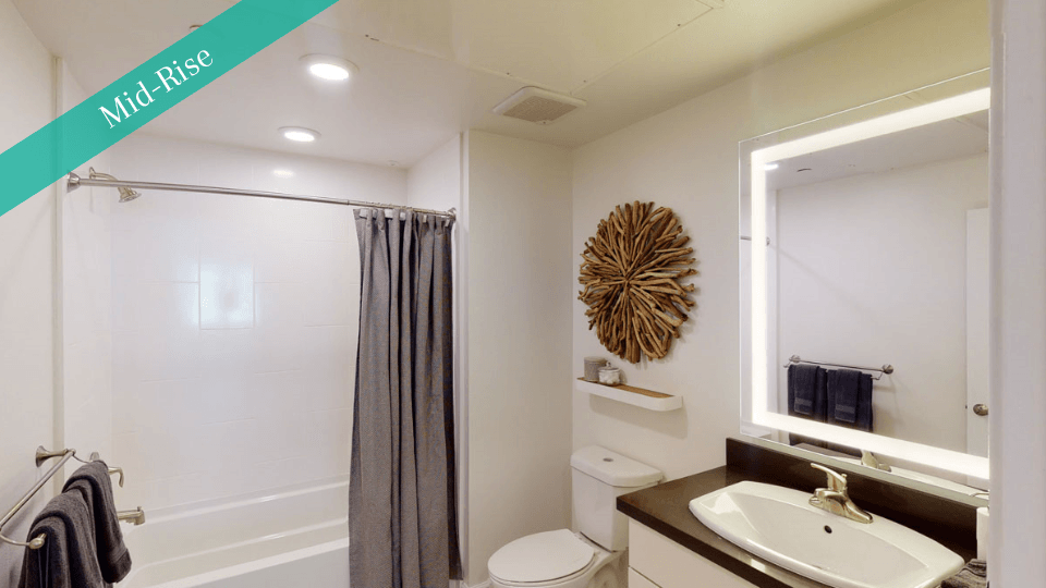 Modern bathroom here at Capitol Towers Luxury Midrise Apartments with white walls, a dark countertop, and a lighted mirror. There's a bathtub with a gray curtain and a wooden decorative piece on the wall.