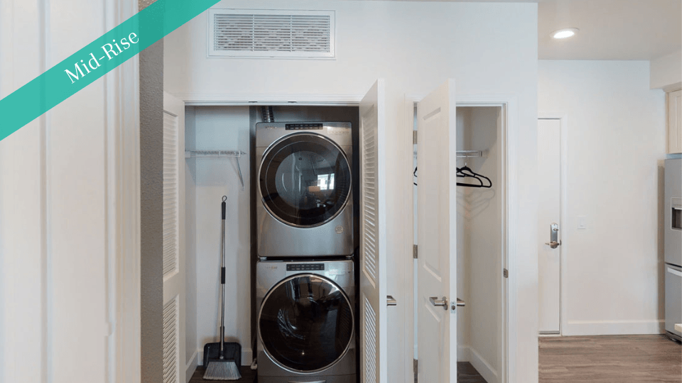 Small laundry area here at Capitol Towers Luxury Midrise Apartments with stacked stainless steel washer and dryer behind open closet doors. Nearby are cleaning supplies and empty hangers.