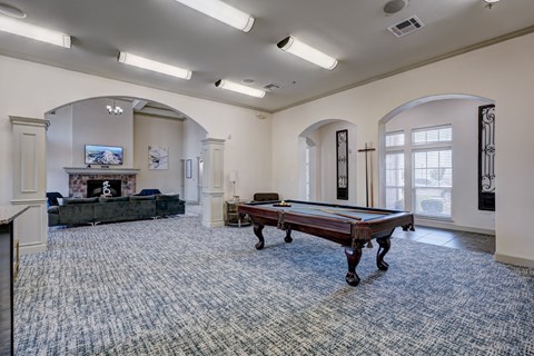 Spacious game room here at Liberty Pointe Apartment Homes with a pool table, surrounded by arched doorways. A cozy seating area with a fireplace and TV is visible in the background. Brightly lit.