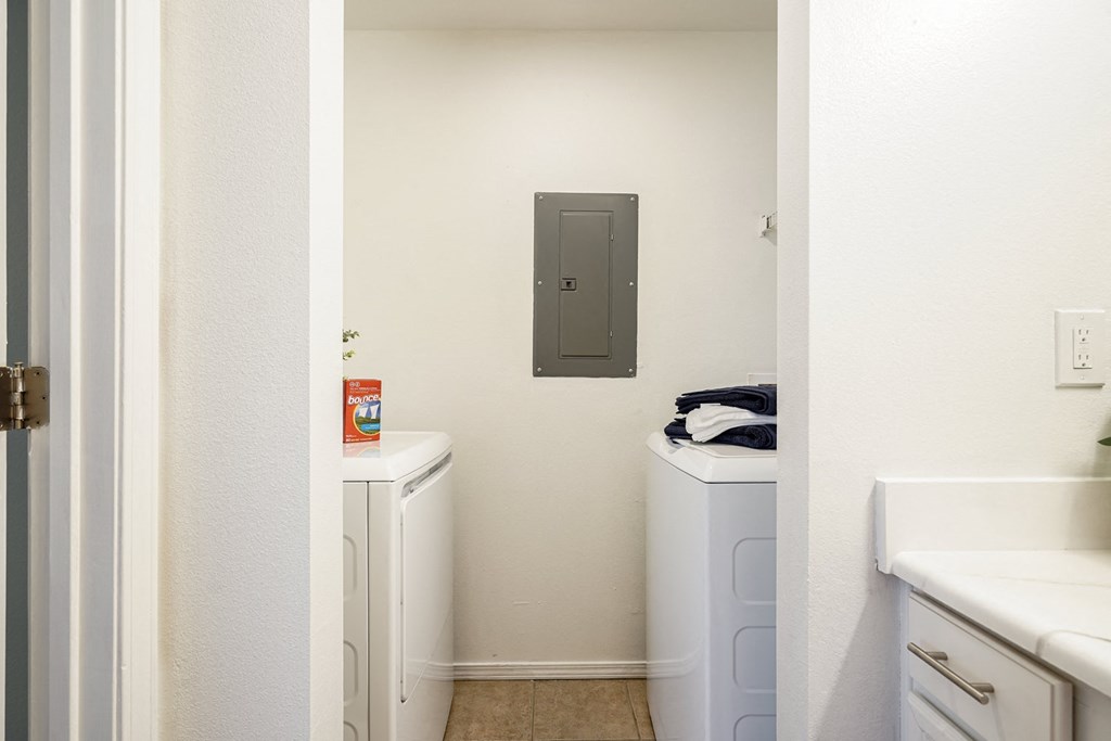 A small laundry room here at Villas at Preston Lakes Apartment Homes with a washer and dryer against a beige wall. A box of dryer sheets sits on the washer, and folded towels are on the dryer.