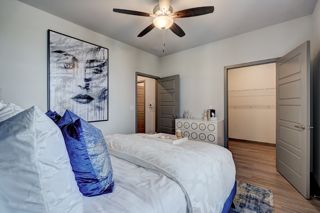 Bedroom here at 35 Degrees North Apartment Homes with a white bed, blue pillows, abstract portrait art, and ceiling fan. Open closet and modern dresser in the background; calm, modern vibe.