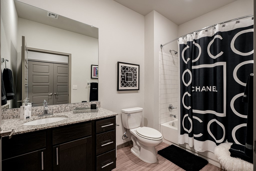 Modern bathroom here at 35 Degrees North Apartment Homes with a granite-topped vanity, large mirror, and white toilet. A black-and-white Chanel-themed shower curtain adds elegance.