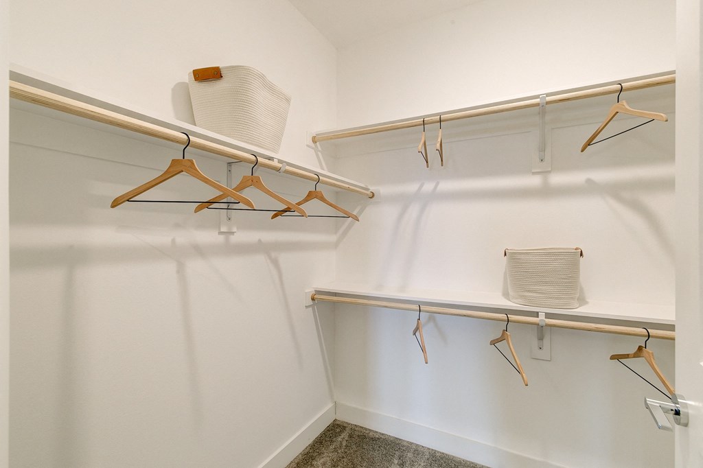 Minimalist walk-in closet here at Florence at the Harbor Apartment Homes with white walls, wooden hangers on rods, and woven baskets on shelves. The space is clean and organized.