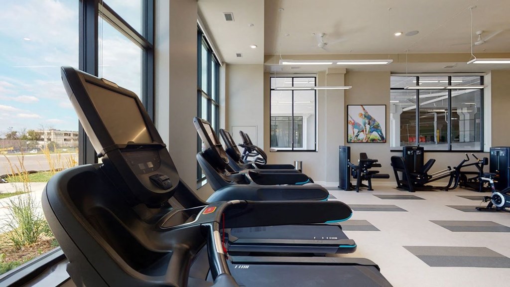 Modern gym interior here at The Collection at Highland Bridge Apartment Homes with several treadmills by large windows, allowing natural light. Neutral tones create a calm, inviting atmosphere for exercise.