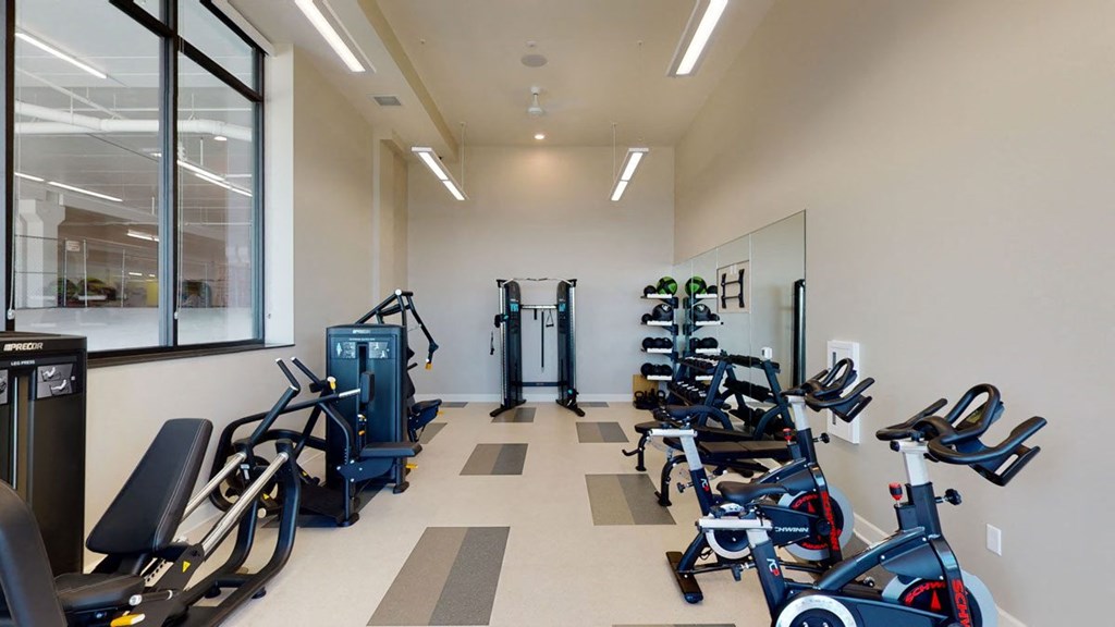 Modern gym here at The Collection at Highland Bridge Apartment Homes with exercise machines, indoor bikes, weight racks, and a mirrored wall. Bright lighting and beige walls create a clean, inviting space.