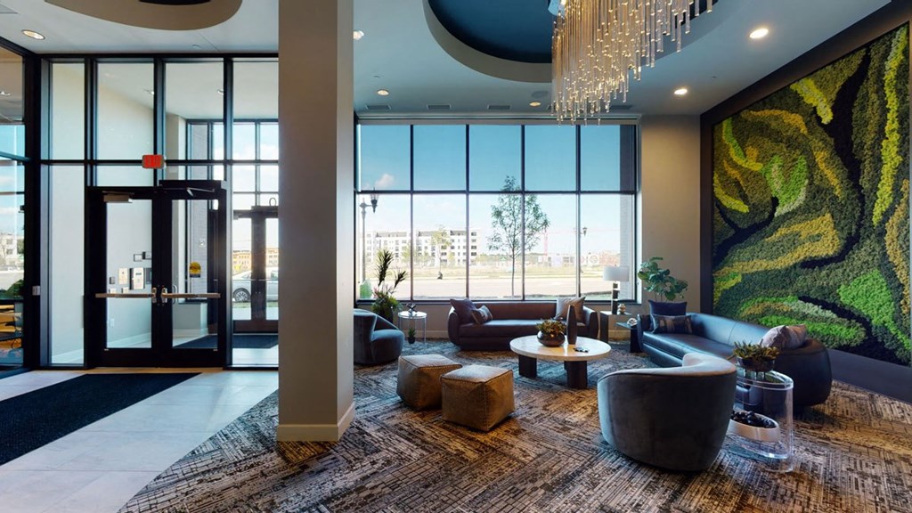 Modern lobby here at The Collection at Highland Bridge Apartment Homes with large windows, plush seating, and vibrant green wall art. A chandelier adds elegance, creating a welcoming atmosphere.
