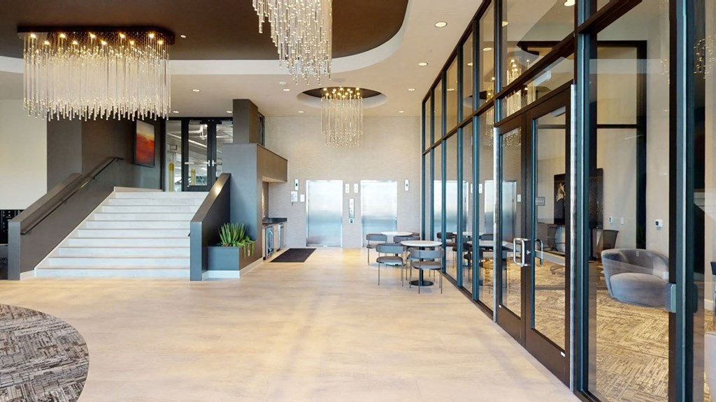 Modern office lobby here at The Collection at Highland Bridge Apartment Homes with glass walls, elegant chandeliers, and sleek furniture. A staircase leads to an upper floor, conveying a sophisticated ambiance.