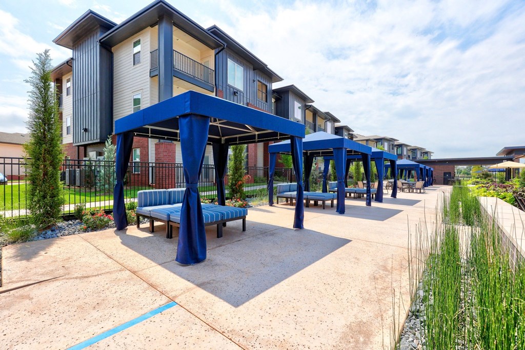 Outdoor scene here at Trailwinds Apartment Homes with blue cabanas lined up on a spacious patio, alongside modern multi-story buildings. Greenery and seating create a relaxed, inviting ambiance.