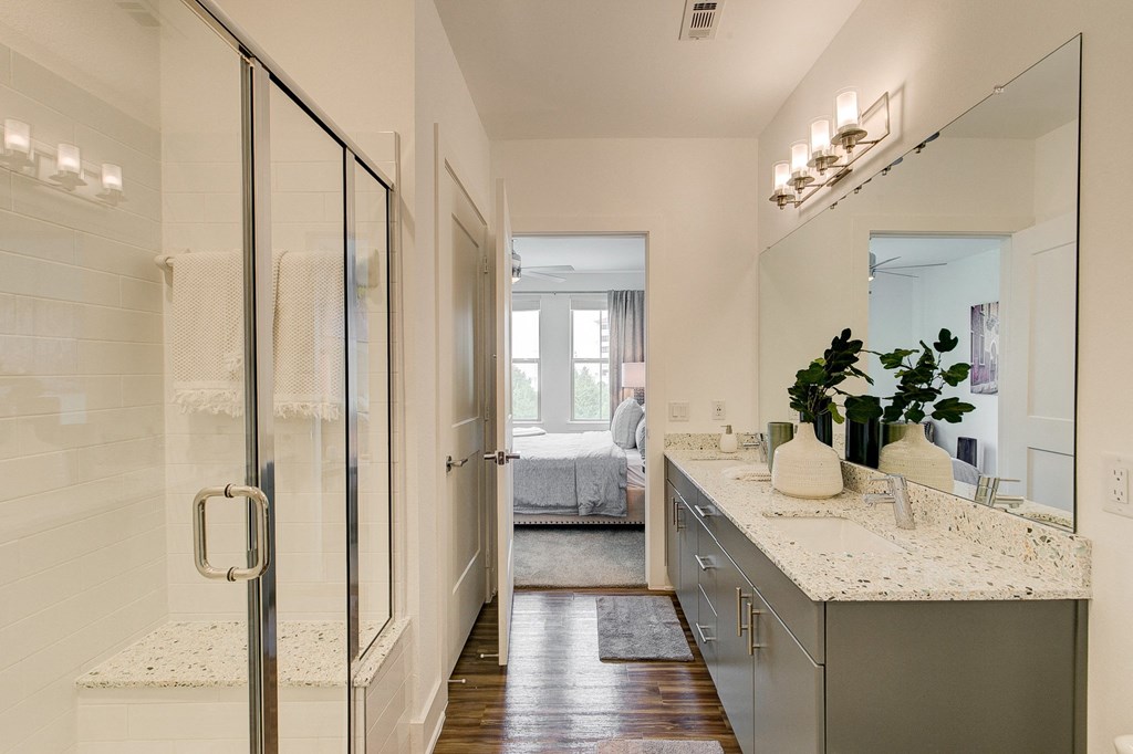 Modern bathroom here at Florence at the Harbor Apartment Homes with a glass shower on the left, gray vanity with a speckled countertop, and decorative vases on the right. Door leads to a bright bedroom.