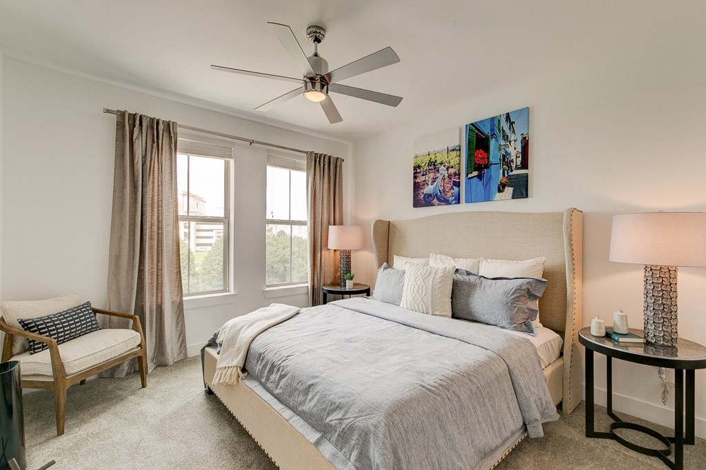 A serene bedroom here at Florence at the Harbor Apartment Homes with a soft beige bed, gray bedding, and cushioned pillows. Art above the bed, a ceiling fan, soft lighting, and cozy accents create a calming atmosphere.