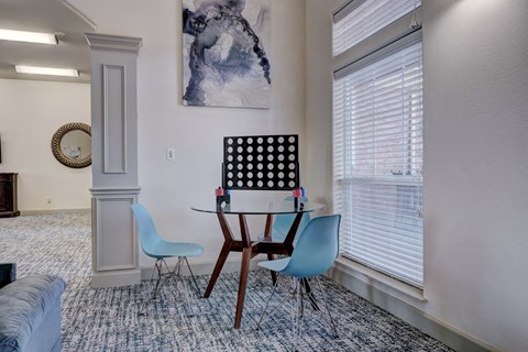 A modern game room here at Liberty Pointe Apartment Homes with a glass table, two blue chairs, and a Connect Four game. A large abstract painting hangs on the wall, and sunlight streams through the blinds.