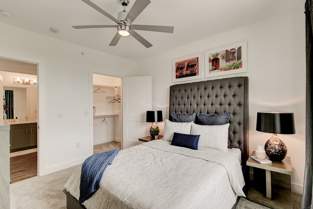 Modern bedroom here at Florence at the Harbor Apartment Homes with a gray upholstered bed, navy and white bedding, and two dark table lamps on nightstands. Open closet and bathroom entrance visible. Bright, cozy atmosphere.