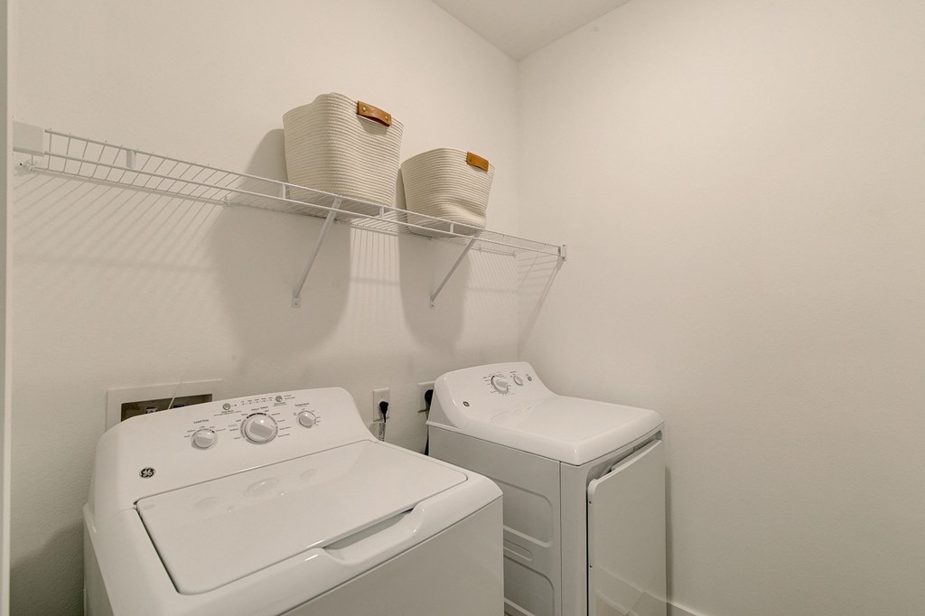Laundry room here at Florence at the Harbor Apartment Homes with white walls features a washing machine and dryer side by side. Above, two beige baskets sit on a wire shelf. Clean and minimal design.