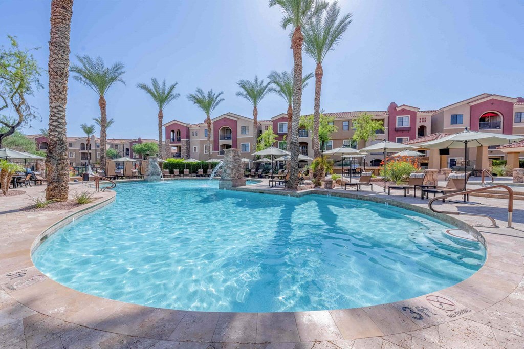 Luxurious pool area here at Desert Club Apartments with clear blue water surrounded by palm trees and sun loungers. Mediterranean-style apartments in the background under a bright sky.