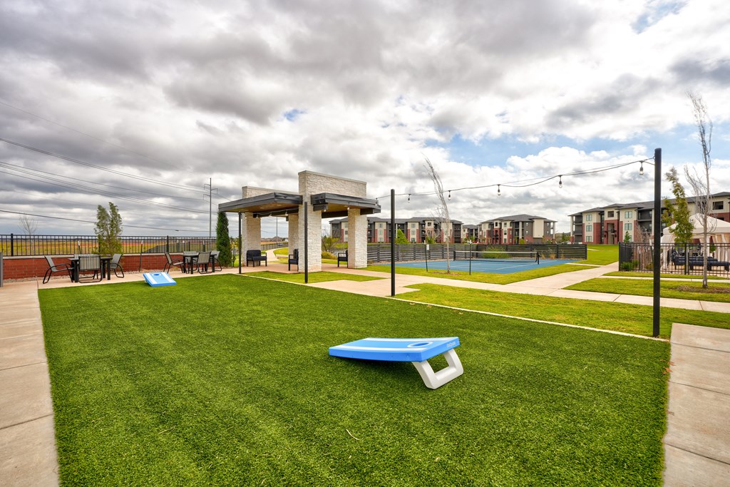 Outdoor recreational area here at 35 Degrees North Apartment Homes with a cornhole game on green grass, white pergola with seating, tennis court in background, overcast sky and string lights.