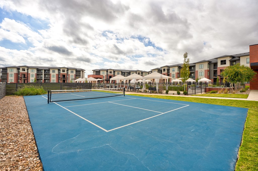 A blue tennis court here at 35 Degrees North Apartment Homes under a partly cloudy sky with modern apartment buildings in the background. Nearby, white canopies cover outdoor seating areas.