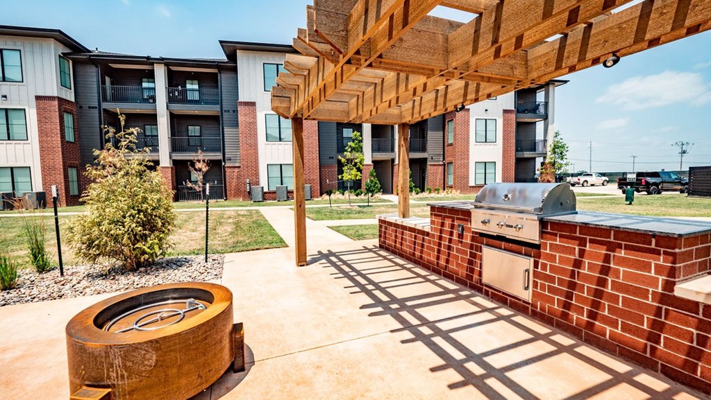 Outdoor BBQ area here at 35 Degrees North Apartment Homes with a brick grill under a wooden pergola. Nearby is a modern fire pit. The background shows a multi-story apartment building.