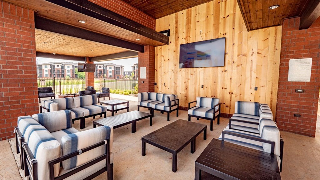 Covered patio here at 35 Degrees North Apartment Homes with striped cushioned seating, wooden ceiling, and brick walls. A large TV is mounted on the wooden wall. Apartments are visible outside.