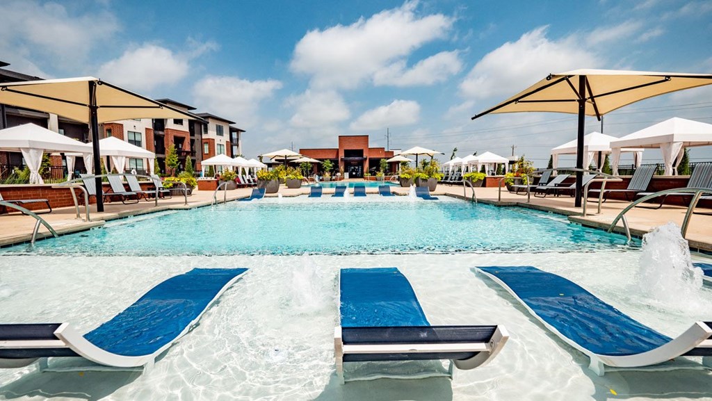 Luxurious outdoor pool here at 35 Degrees North Apartment Homes with cascading water features, blue lounge chairs, and large umbrellas. Modern apartments and cabanas frame the vibrant scene.