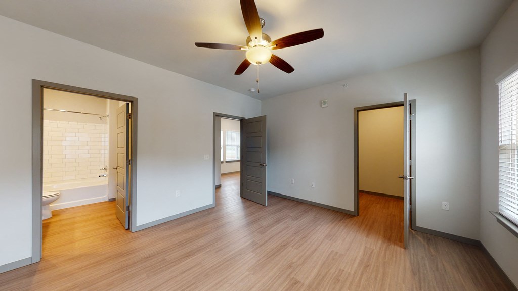 Empty room here at 35 Degrees North Apartment Homes with wood floors, gray doors, and a ceiling fan. Open doors reveal a bright bathroom with white tiles and a window-lit adjacent room.