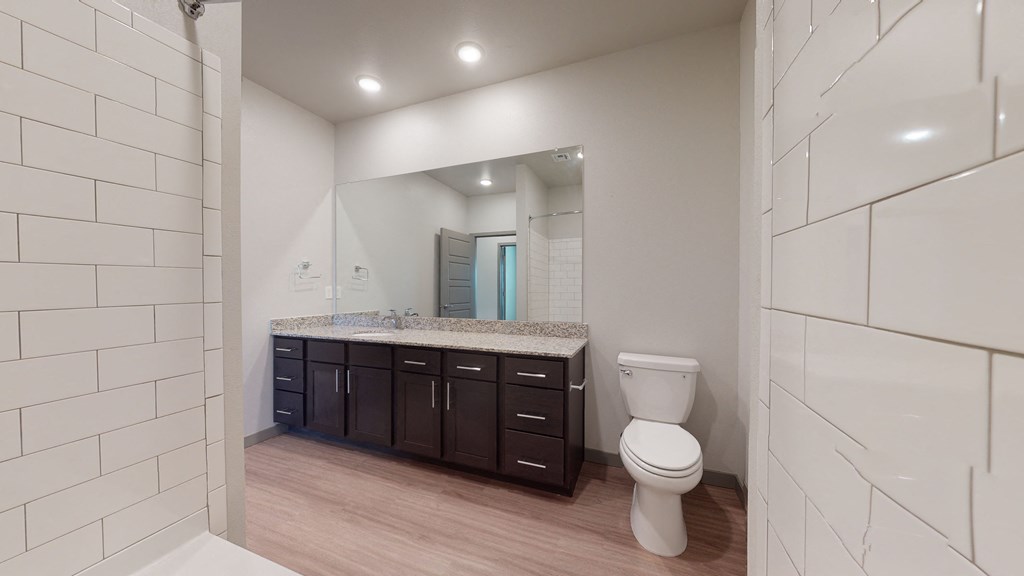 Modern bathroom here at 35 Degrees North Apartment Homes with white subway tile, large mirror, granite countertop, dark wood cabinets, and a white toilet. Calm and clean ambiance.