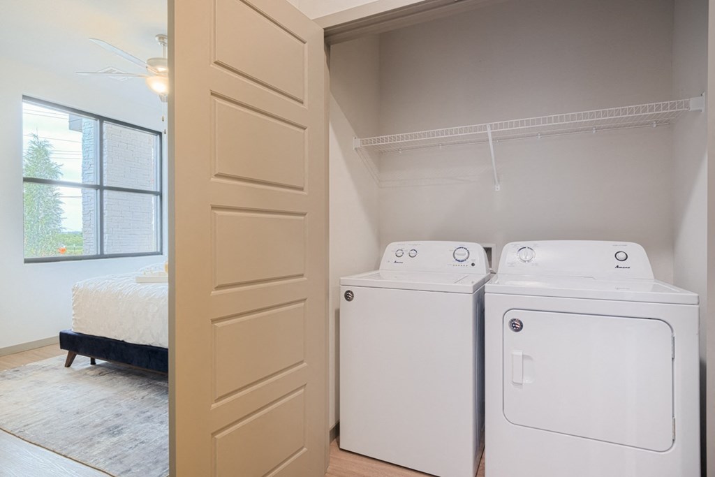 Laundry area here at 35 Degrees North Apartment Homes with a white washer and dryer in a closet. Beige sliding door partially open, revealing a bright bedroom with a bed and large window. Calm and tidy atmosphere.