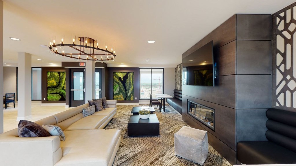 Modern lounge here at The Collection at Highland Bridge Apartment Homes with beige sectional sofa, geometric chandelier, and wall-mounted TV. Textured rug and plant artworks add a stylish, cozy touch.