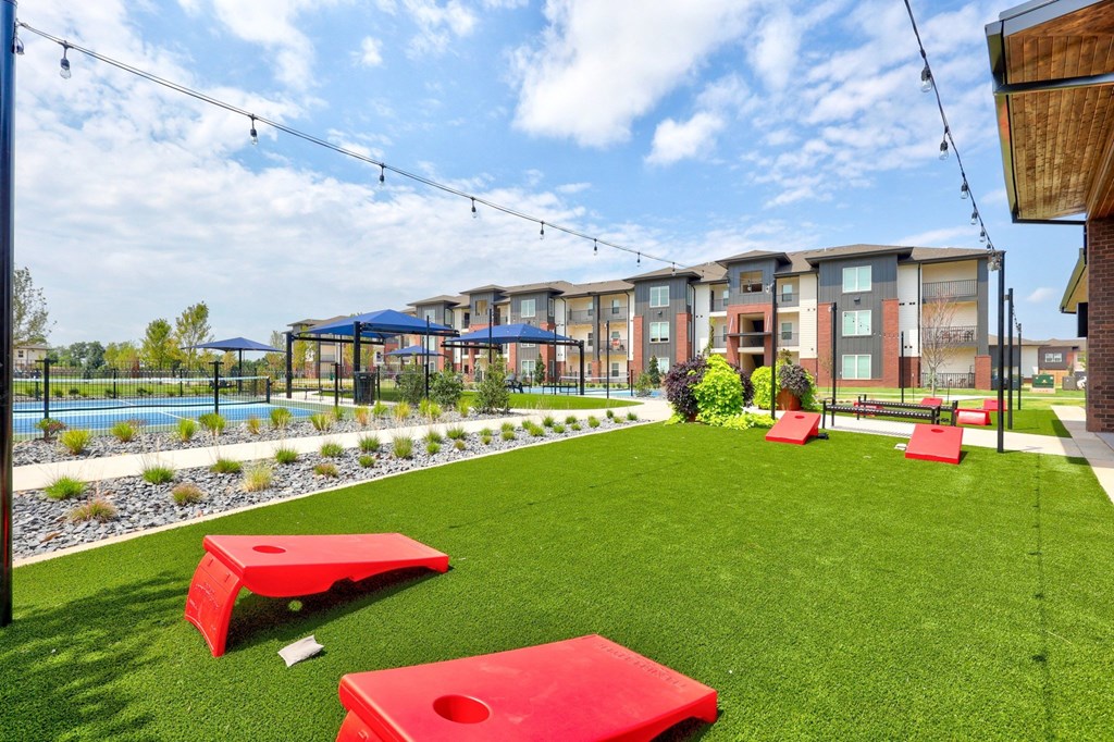 Trailwinds Apartment Homes with red cornhole boards on green grass, string lights above, and blue umbrellas near a pool. Bright, clear day with a relaxed vibe.