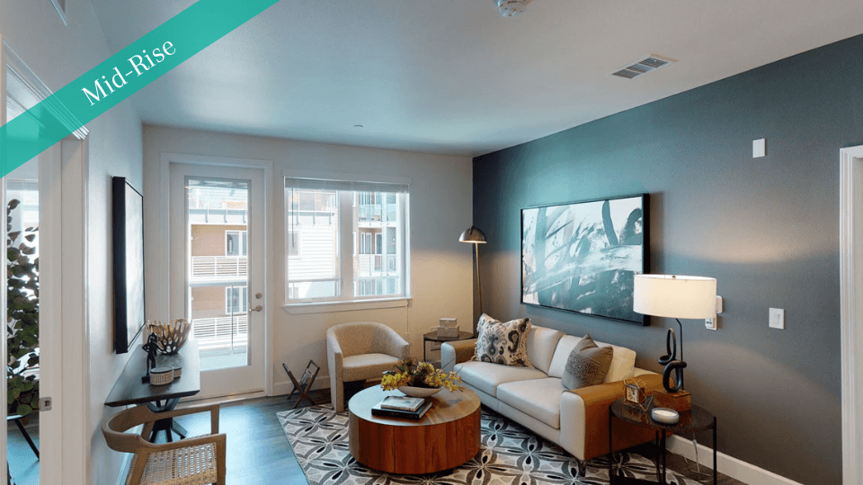 A modern living room here at Capitol Towers Luxury Midrise Apartments with a sleek gray accent wall, a beige sofa adorned with patterned pillows, a round wooden coffee table, and abstract wall art. A large window and door let in natural light, creating a cozy ambiance. A "Mid-Rise" label is in the top left corner.