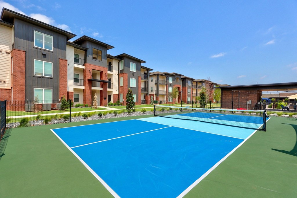 Modern Trailwinds Apartment Homes with brick and beige siding under a clear blue sky. A blue pickleball court in the foreground adds a recreational touch.