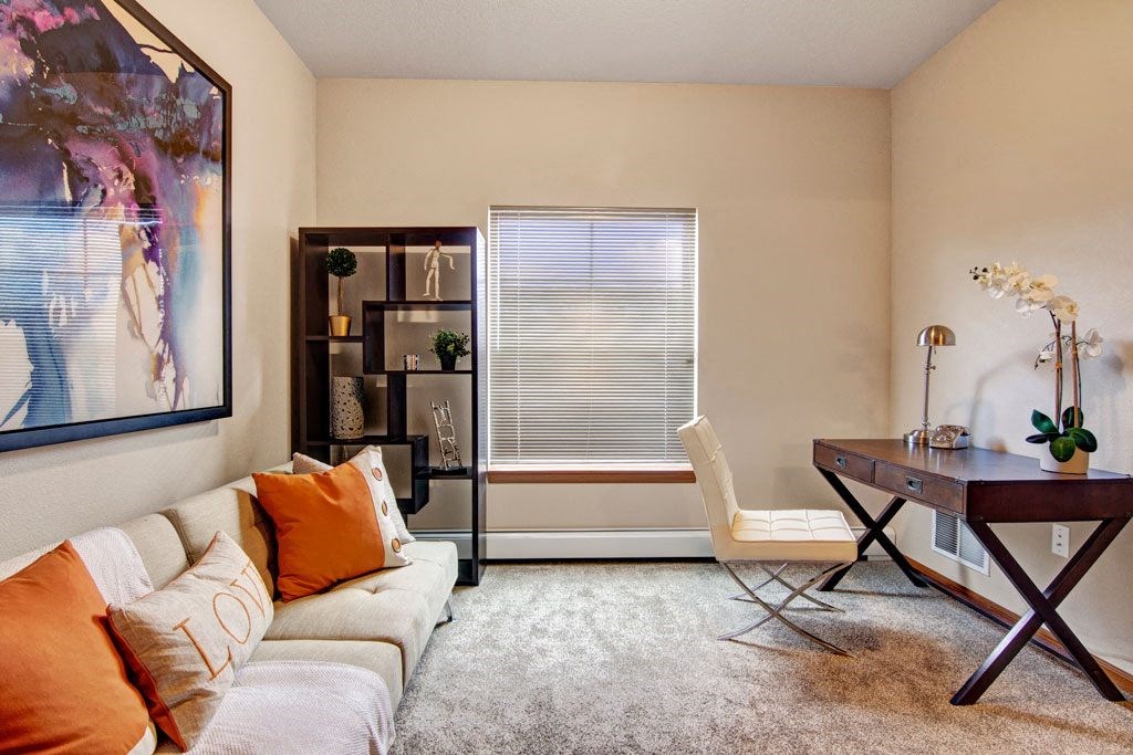 A modern home office here at The Club at Heritage Apartment Homes with a beige sofa adorned with orange pillows, a geometric bookshelf, abstract wall art, a sleek dark desk, and a window with blinds.
