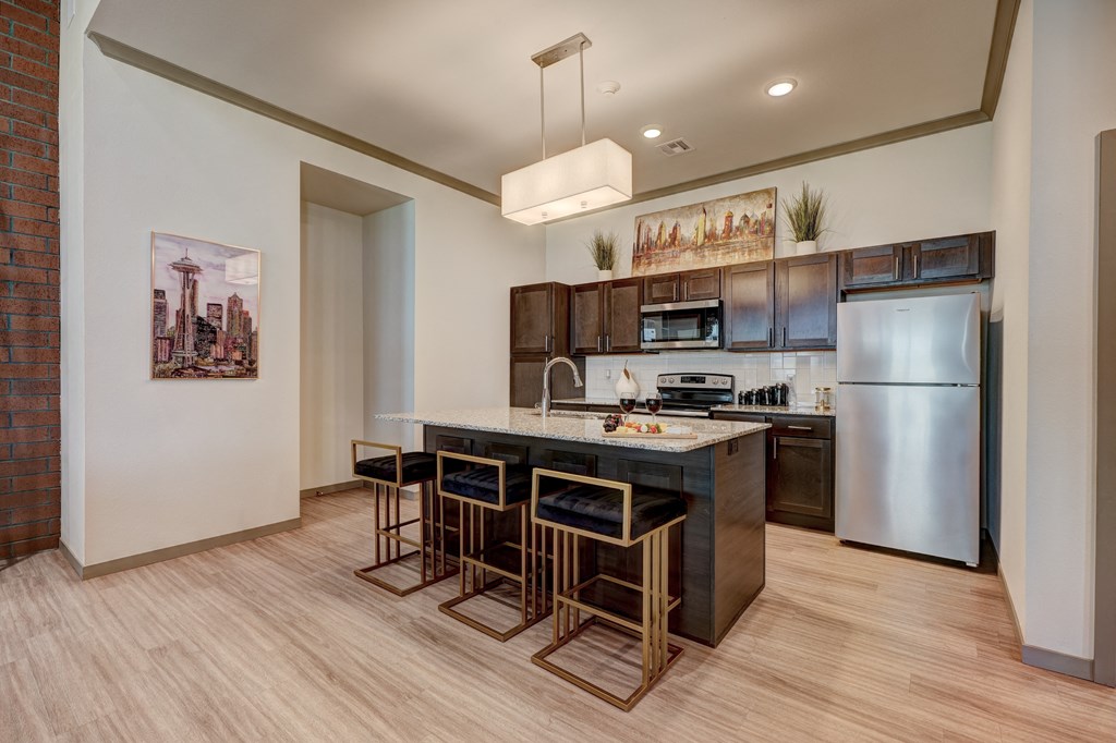 Modern kitchen here at 35 Degrees North Apartment Homes with dark wood cabinets and a granite island. Four sleek stools surround the island, with wine glasses and a snack on top. Warm lighting and art enhance the cozy ambiance.
