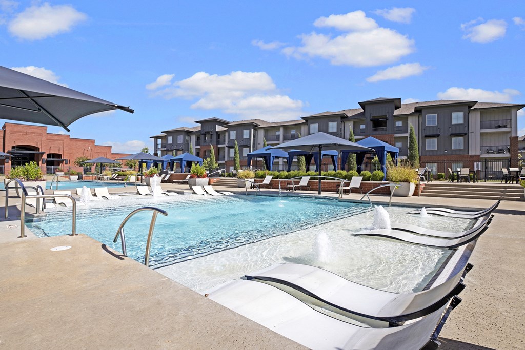 Outdoor pool area here at 71 at Tulsa Hills Apartment Homes with lounge chairs and water fountains under a clear blue sky. Modern apartments surround the area, creating a relaxing ambiance.
