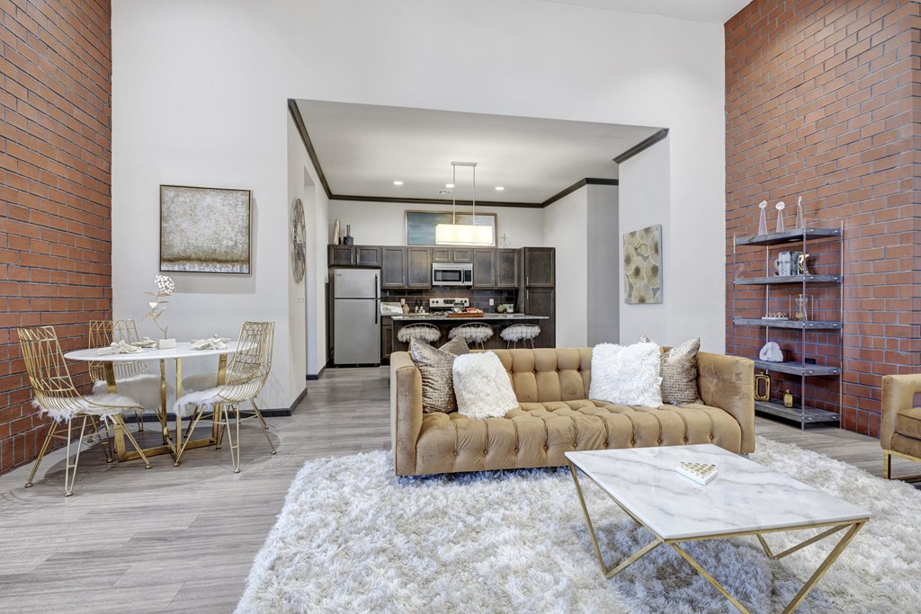 Modern seating area here at 71 at Tulsa Hills Apartment Homes with a tan sofa, fluffy white rug, and marble coffee table. Exposed brick, gold accents, and an open kitchen create a warm, chic atmosphere.