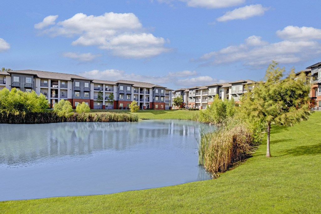 71 at Tulsa Hills Apartment Homes complex with pond and landscaped greenery under a blue sky with scattered clouds. The serene setting conveys a peaceful, suburban vibe.