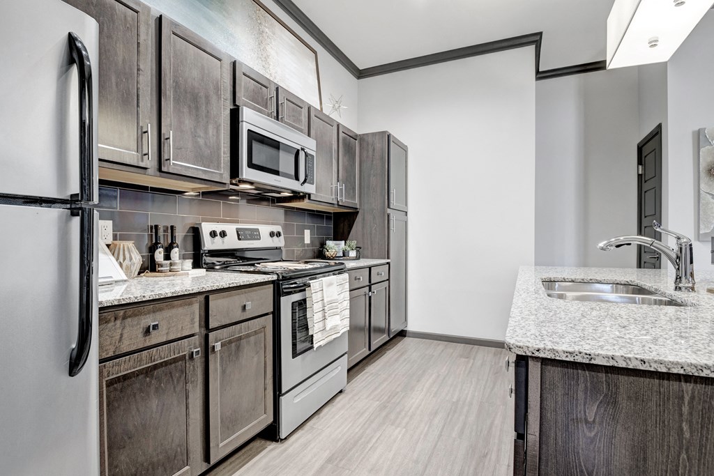 Modern kitchen here at 71 at Tulsa Hills Apartment Homes with dark wooden cabinets, granite countertops, and stainless steel appliances. A cozy and clean atmosphere with a sleek design.