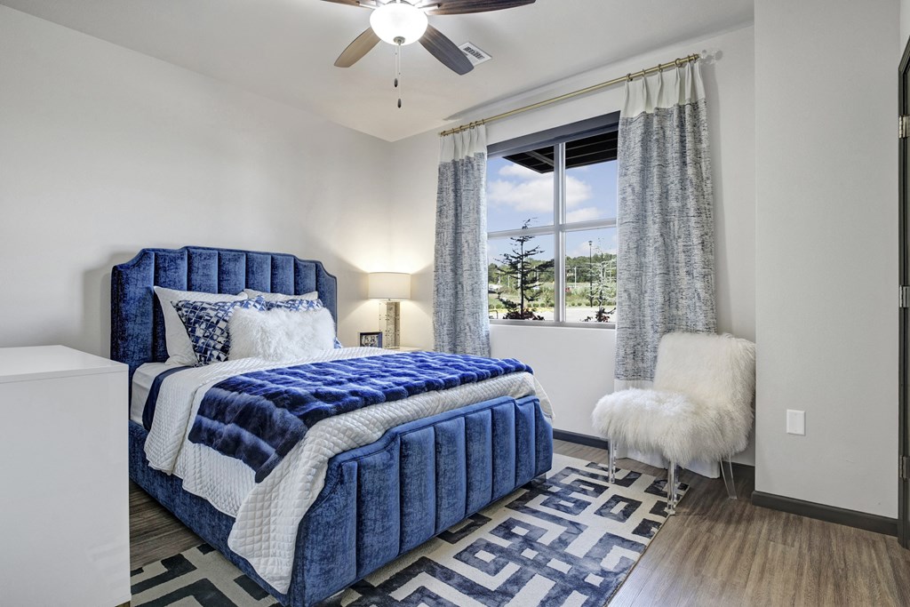 Bright bedroom here at 71 at Tulsa Hills Apartment Homes with a plush blue bed, white and blue bedding, and a geometric rug. A fluffy white chair is beside a window with scenic views.