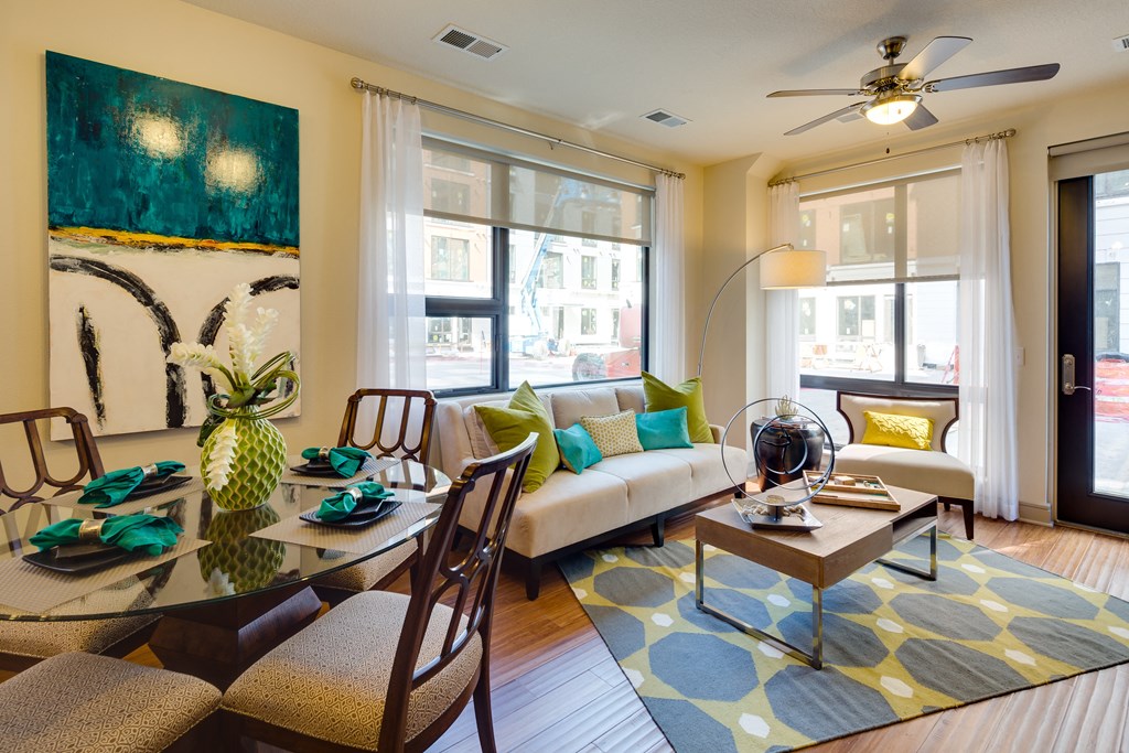 Modern living room here at Inspire Apartment Homes with a beige sofa adorned with green and turquoise pillows, abstract art, a glass dining table, and a geometric rug. Bright and inviting.