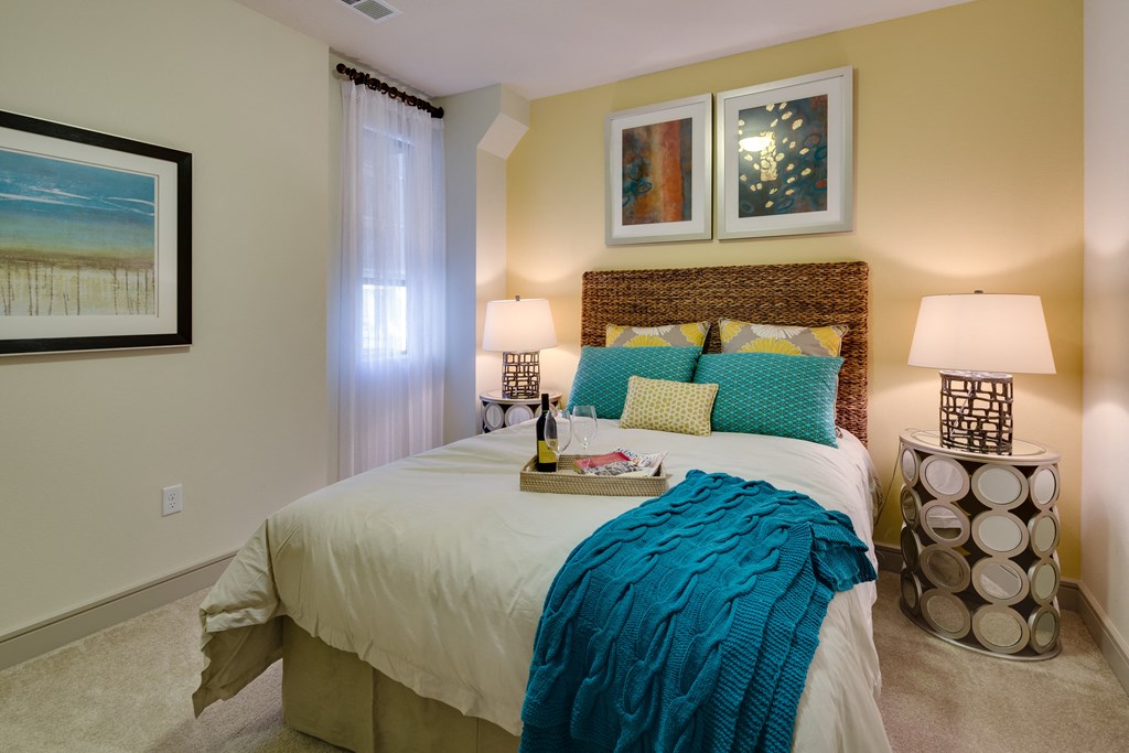 Cozy bedroom here at Inspire Apartment Homes with a wicker headboard, teal bedding, and yellow accents. Soft lighting from modern lamps, abstract art, and a tray with wine add elegance.