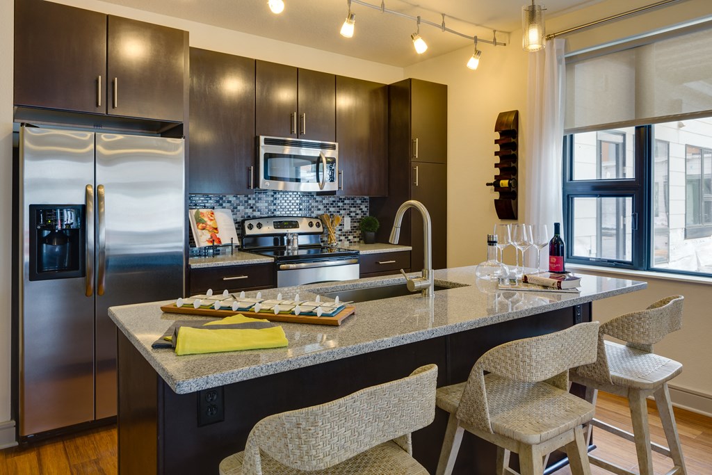 Modern kitchen here at Inspire Apartment Homes with dark wood cabinets, stainless steel appliances, and a granite island with wicker stools. Warm lighting and a cozy ambiance.