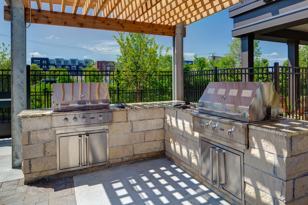 Outdoor patio here at Inspire Apartment Homes with two stainless steel grills on a stone counter under a wooden pergola. Shadow patterns on the ground and sunny greenery in the background.
