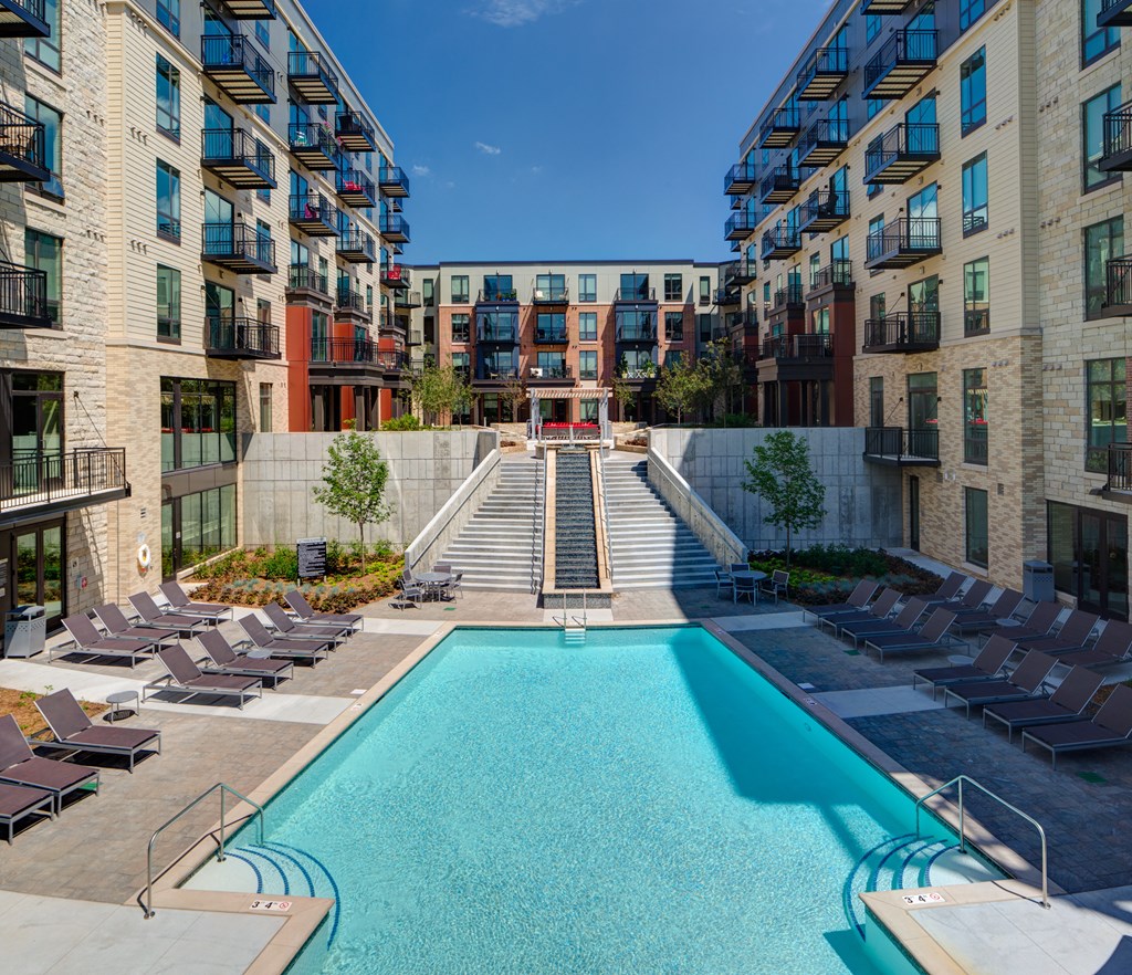 Modern Inspire Apartment Homes complex with a central pool and surrounding sun loungers. Flanked by two five-story buildings, a staircase leads to a rooftop terrace.